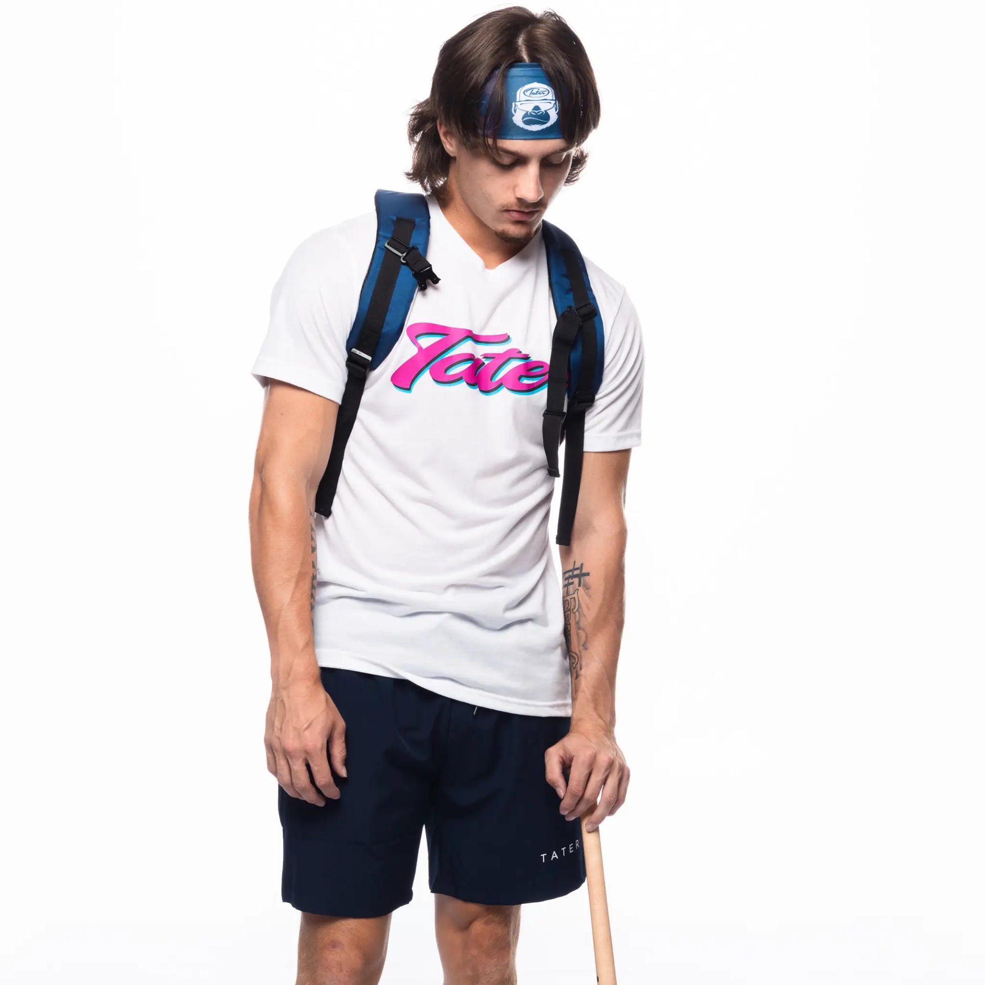 In the image, there is a male model wearing a white T-shirt with a colorful "Tater" logo on the front, navy workout shorts with the Tater brand name, a baseball headband, and a black backpack. He is also holding a wooden baseball bat in his right hand. The overall look appears to be casual and suitable for both athletic activities and casual wear.