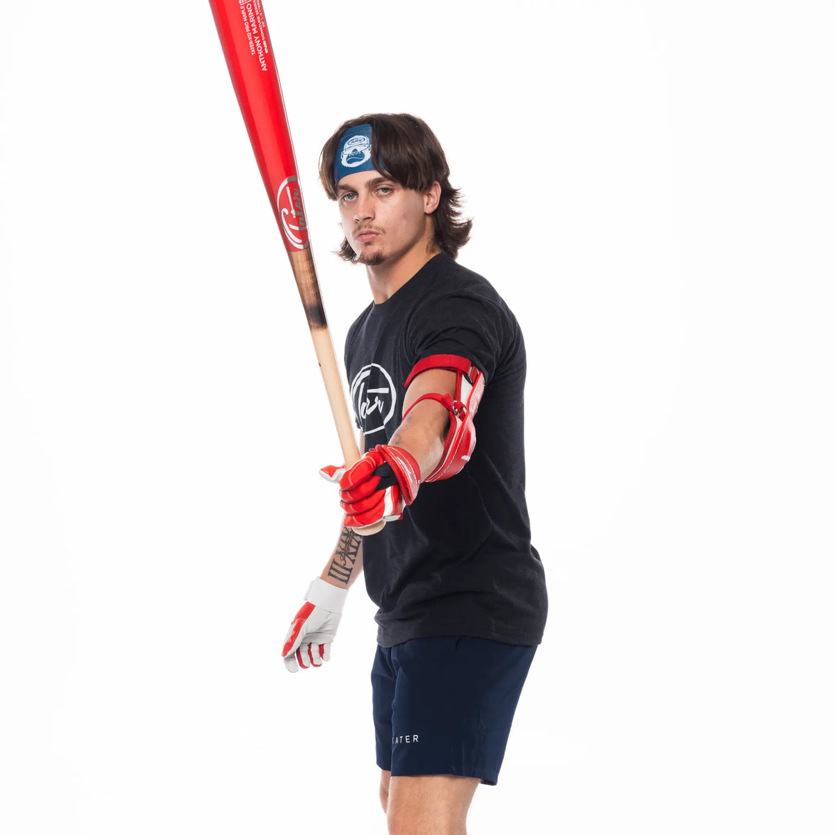The image features a baseball player posed with a bat over his shoulder. He is wearing a black T-shirt with the Tater Baseball logo, navy workout shorts also branded with the Tater logo, and red-and-white batting gloves. The outfit is a sporty, cohesive look suitable for baseball training and casual wear.