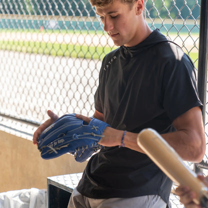 A baseball player is seen wearing a black short-sleeve BP (batting practice) hoodie from Tater Baseball. He is holding a blue outfielder's glove and a natural wood bat, indicating he's possibly preparing for practice or a game.