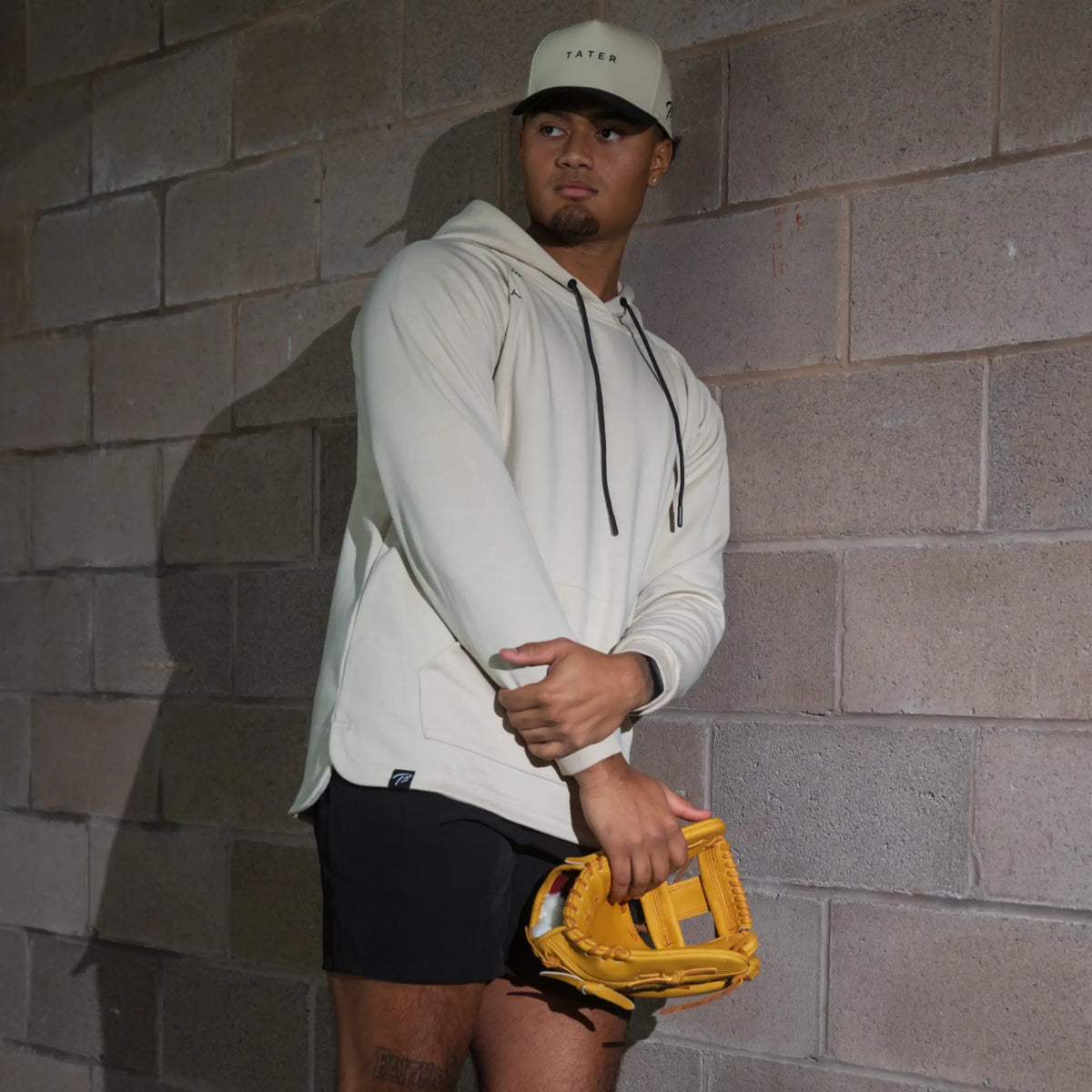 In the photo, a baseball player is wearing a cream-colored TATER-branded hoodie and cap, with a yellow infielder&#39;s glove, showcasing athletic wear suitable for both sports and leisure.