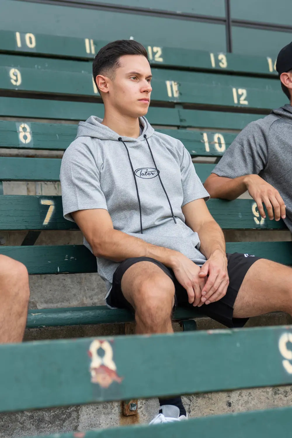 Baseball scout sitting on the bleachers wearing Tater athletic hoodie and shorts while watching players practice at Muzzy Field in Bristol, Connecticut.