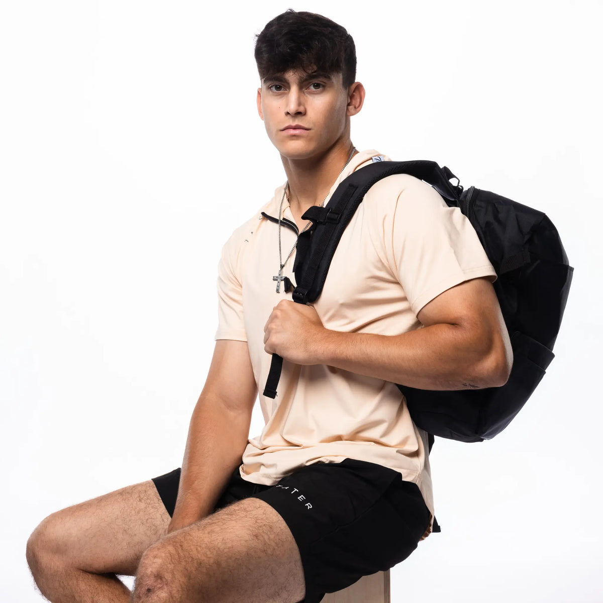 In the image, there is a young man wearing a tan short-sleeved hoodie and black athletic shorts, both featuring the &quot;TATER&quot; logo, which suggests they are part of a baseball athletic wear line. He is also carrying a black backpack over one shoulder and has a set of earphones around his neck. His attire and accessories indicate a casual, sporty aesthetic, likely targeting athletes or individuals with an active lifestyle.