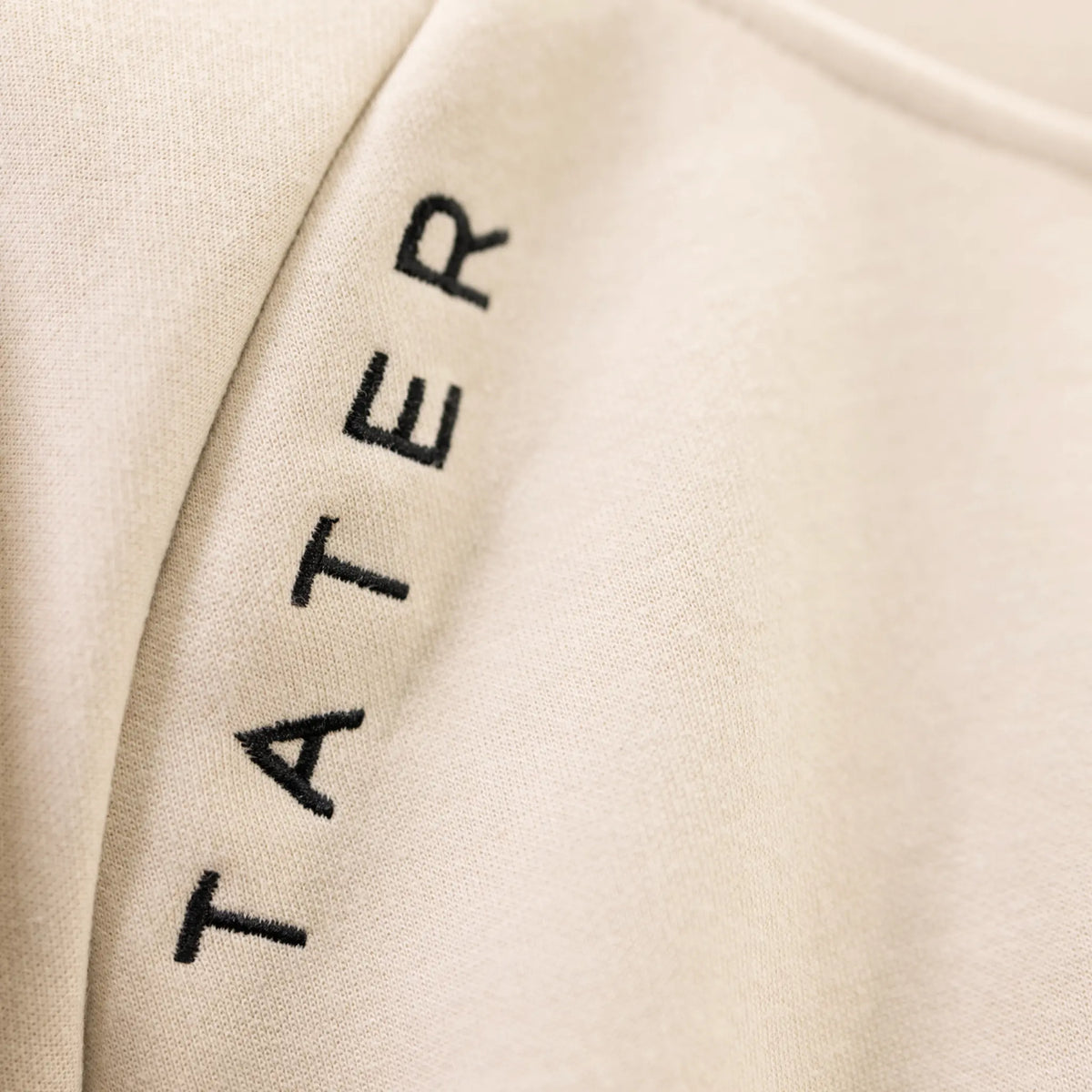The image shows a close-up of a Tater Baseball branded workout hoodie in a cream color. The TATER logo is embroidered in black, emphasizing the premium material of the apparel.