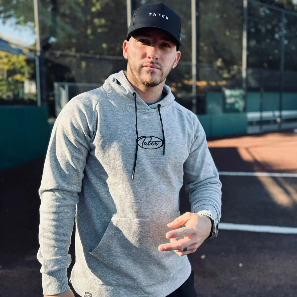 Confident man posing in a Tater Baseball athleisure grey hoodie with long sleeves, paired with a black Tater cap, exemplifying a casual, sporty style suitable for both training and everyday wear.
