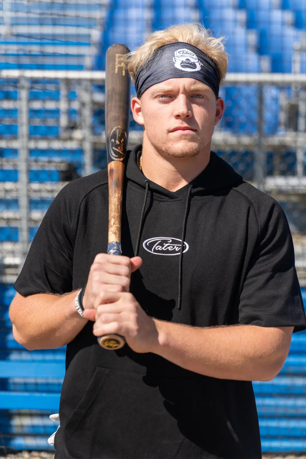 Benjamin Huber wearing a black mens baseball hoodie and headband. He is gripping a 32 inch maple wood bat while standing in front of the Municipal Stadium bleachers.